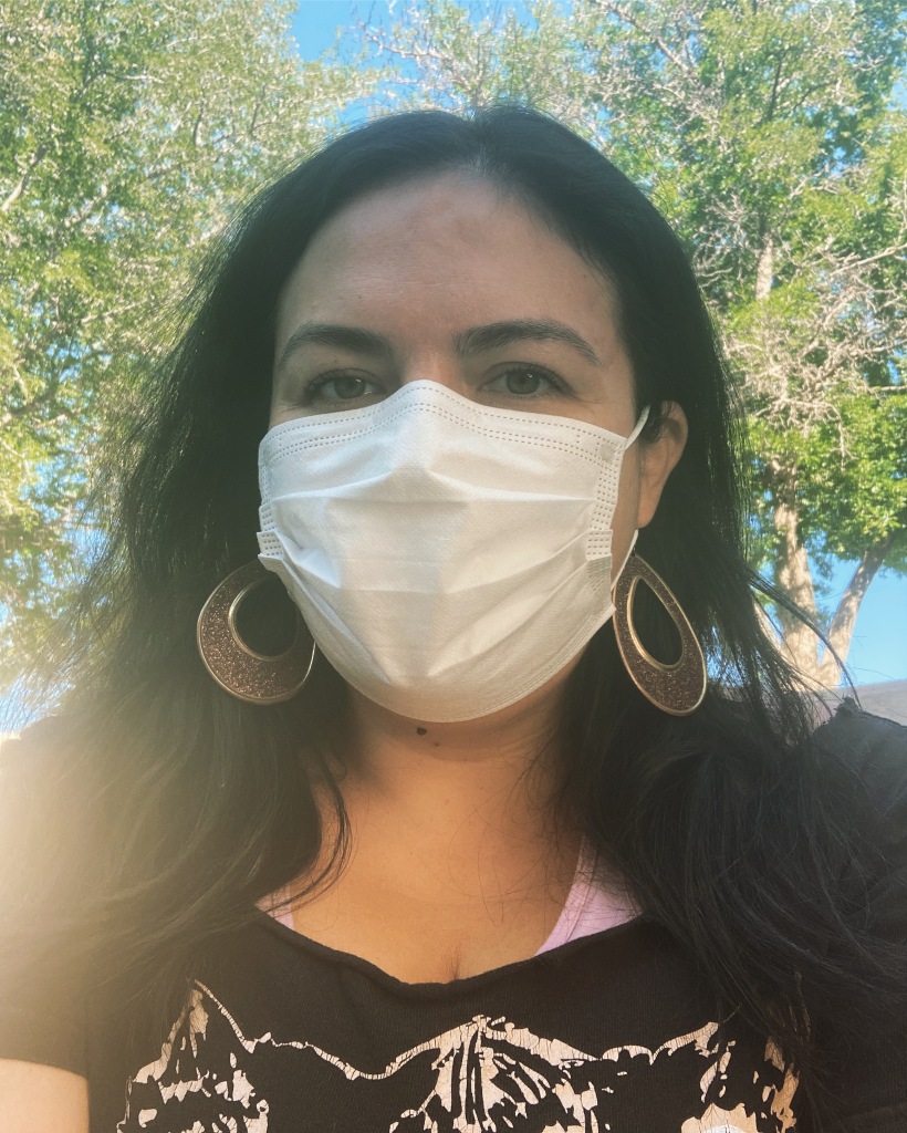 Alisha is looking at the camera and wearing a white surgical mask, glittery teardrop earrings, and a black shirt with her black hair set against green trees in the background. 